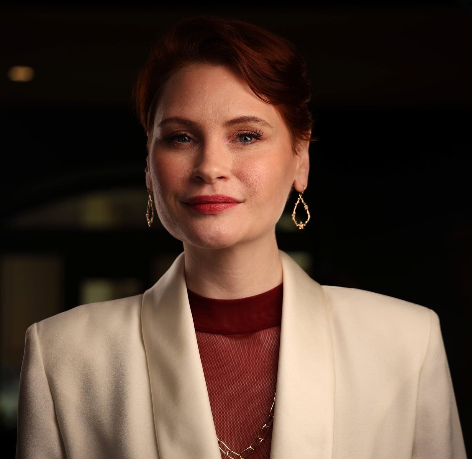 Headshot in a cream blazer and gold earrings against a dark background