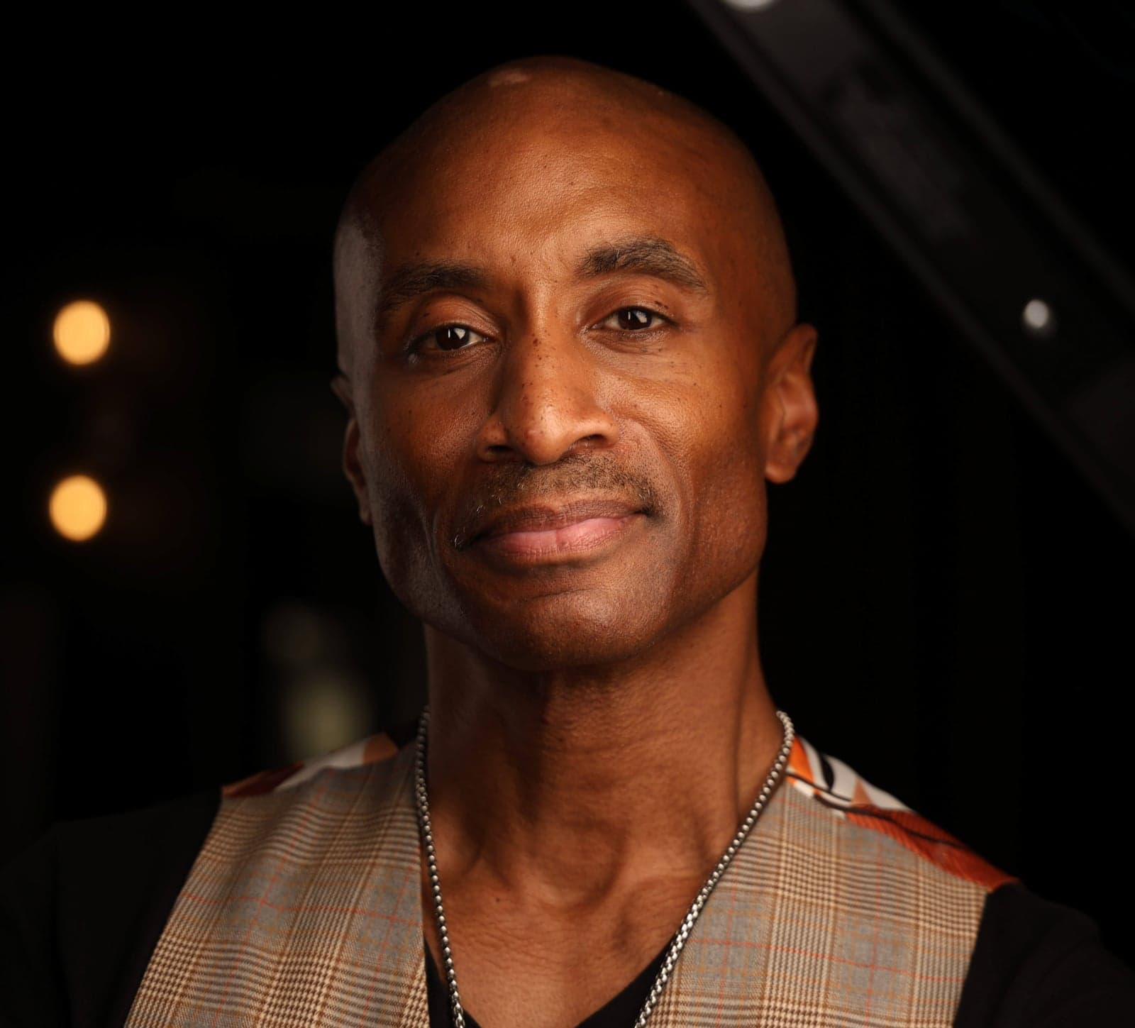 Headshot on a dark background with warm bokeh, plaid vest and chain necklace
